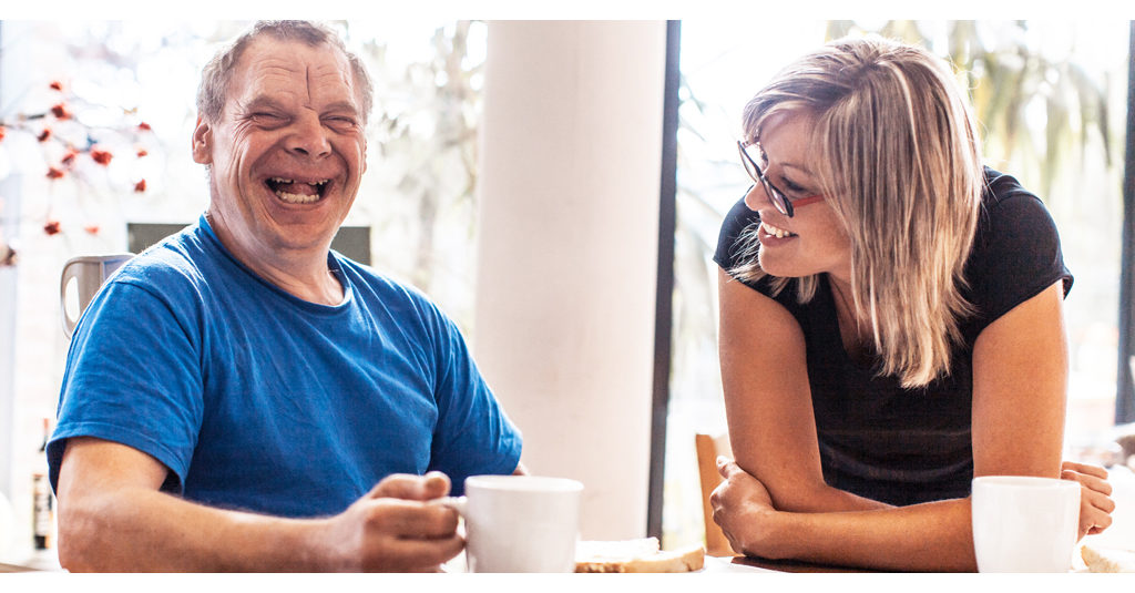 Happy man with a developmental disability sitting at a kitchen table with a woman smiling at him.