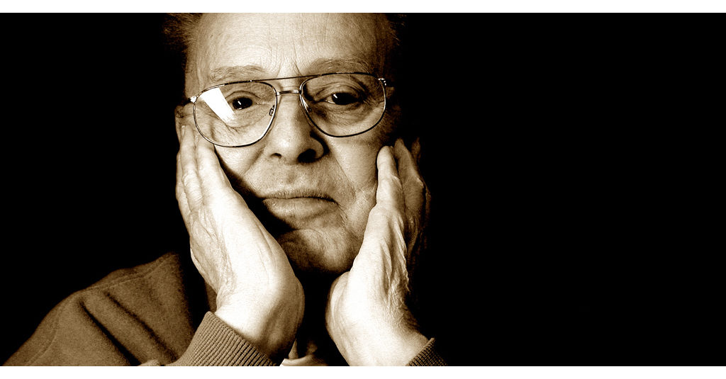 Older man with his hands on his cheeks and a somber stare. Dark background.